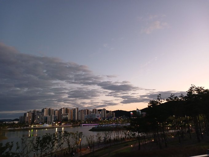 An evening cityscape view across a river showing illuminated high-rise buildings under a twilight sky with scattered clouds.