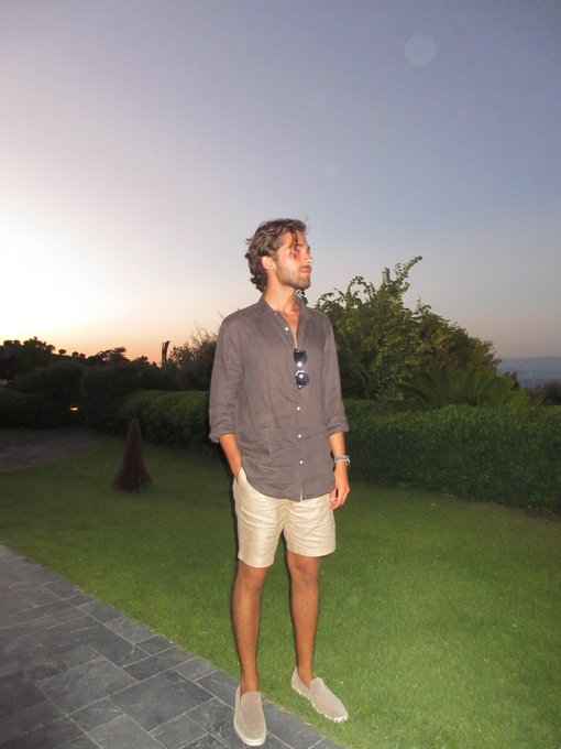 A young man stands on a stone pathway beside a well-manicured lawn at sunset, dressed in a gray button-up shirt, khaki shorts, and light sneakers, with sunglasses tucked into his shirt. The tranquil outdoor setting visually echoes the post's "touch some grass" message, serving as a gentle reminder for crypto enthusiasts to step away from their screens and enjoy the natural world.