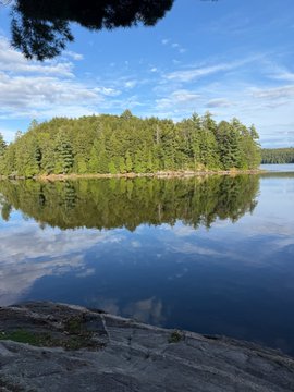 A serene landscape photograph showing a small forested island reflected in a calm lake under a blue sky with scattered clouds.