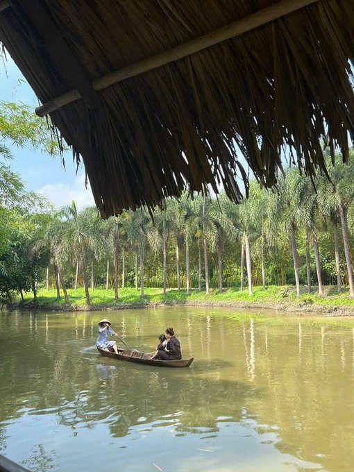 Two people in a small wooden canoe on a calm river lined with palm trees, viewed from beneath a thatched roof structure.