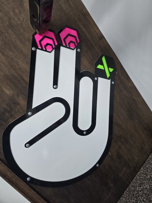 A white foam finger sign with a black border is decorated with pink hexagonal emblems and a green 'X' logo, photographed on a wooden surface.