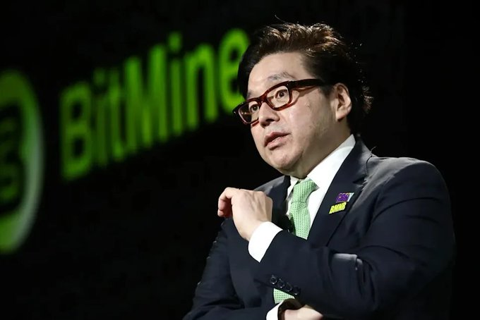 A man in a dark suit and green tie wearing glasses speaks at what appears to be a BitMine event, with the green BitMine logo visible in the background.