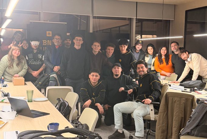 A group of approximately twenty people poses together in what appears to be an office or conference room, with some wearing Raiders apparel and others dressed in casual attire, gathered around tables with laptops and backpacks visible. The "BNB" logo is visible on a display in the background, aligning with the "BNB Builder Sessions BA" caption, suggesting this is a meetup of Binance builders/developers in Buenos Aires.