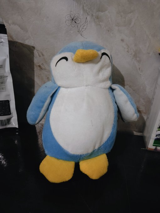 A plush toy of a blue and white penguin with closed smiling eyes, a yellow beak, and yellow feet, sitting on a dark surface.