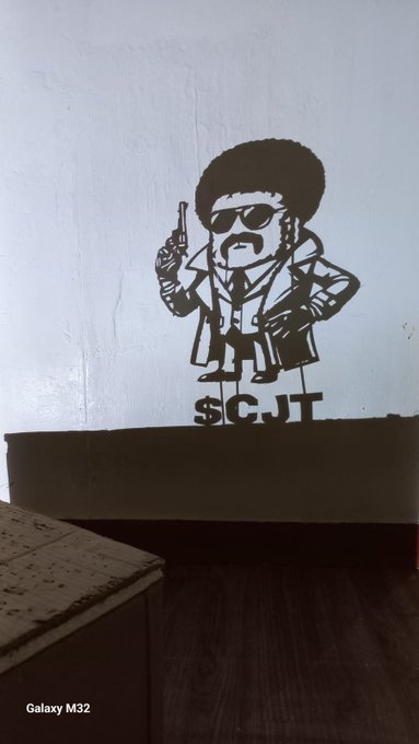 A black stencil graffiti on a white wall depicting a character with an afro, sunglasses, and a trench coat holding a gun, with the text 'SCJT' below.