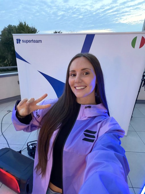 A smiling woman with long dark hair poses in front of a 'superteam' branded backdrop, making a peace sign with her hand while wearing a purple and blue jacket.