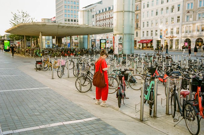 A woman in a red outfit stands amid a crowded bicycle parking area, examining the rows of bikes lined up outside what appears to be a transit station. The scene captures an everyday urban moment of cycling culture, fitting the "Parking Lot" title from the "Northern Observations" series available for 0.069ETH.