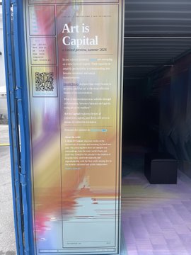 The image shows a poster with a blue frame that has text on it. The poster appears to be advertising an art event or exhibition. It features a photo of a person's body along with some text about the event, which includes details such as the name of the artist and the date of the event. The poster is displayed in front of a building, possibly a gallery or a studio space for artists.