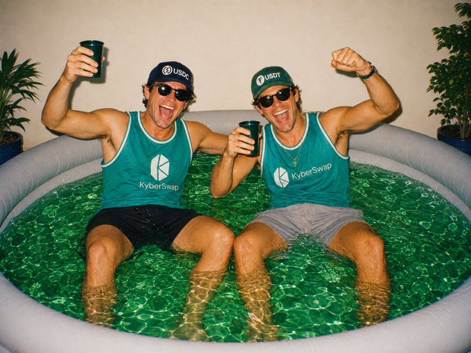 Two men in matching green 'KyberSwap' tank tops and 'USDC' hats sit in an inflatable pool filled with green liquid, raising their fists and cups.