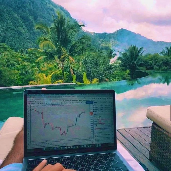 A person uses a MacBook displaying a trading chart while relaxing by an infinity pool overlooking a lush tropical landscape with mountains and palm trees.