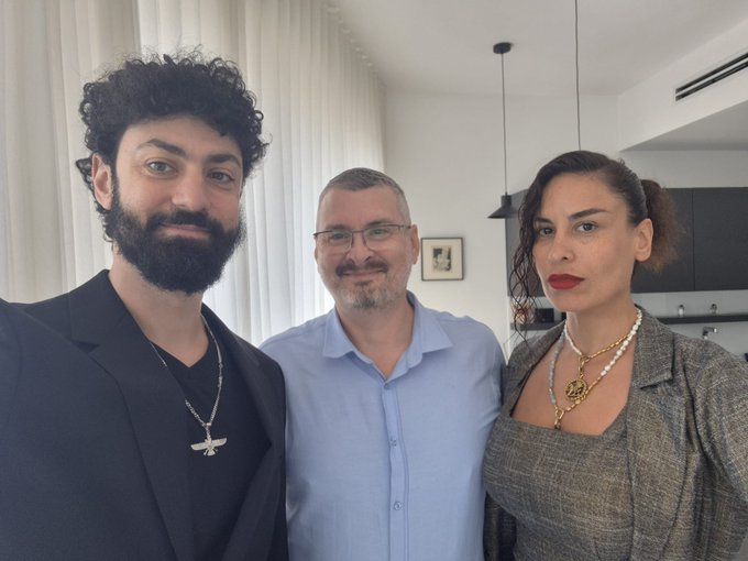 Three people pose for a selfie indoors, with a woman on the right wearing distinctive bright red lipstick, a pearl necklace with a gold pendant, and a grey blazer.