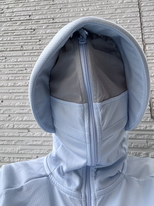 A person wearing a light blue hooded garment with a front zipper and mesh paneling stands against a white brick wall, with the hood pulled up to completely cover their face.