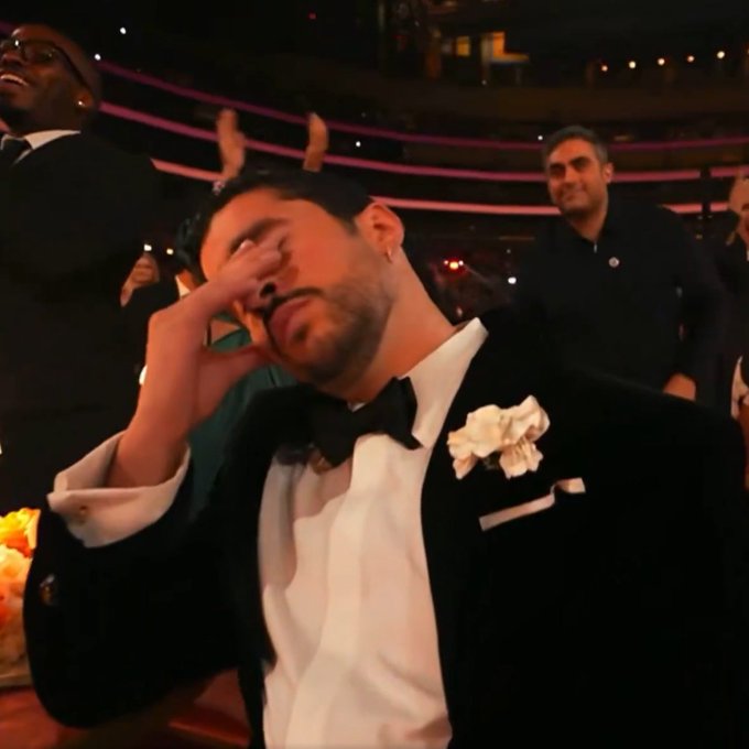 A man in a formal black tuxedo with a white floral boutonnière shields his eyes with one hand while smiling, seated in an audience at what appears to be an awards ceremony.