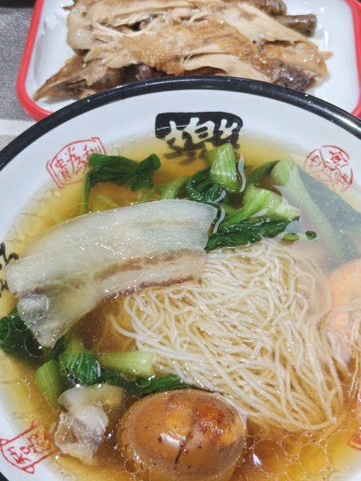 A bowl of ramen with clear broth, noodles, bok choy, chashu pork, and a marinated egg sits beside a plate of shredded chicken, featuring Chinese characters '梅' (plum) on the bowl.