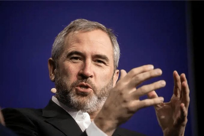 A man with a grey beard and a headset microphone gestures with his hands while speaking at what appears to be a conference, set against a dark blue background.