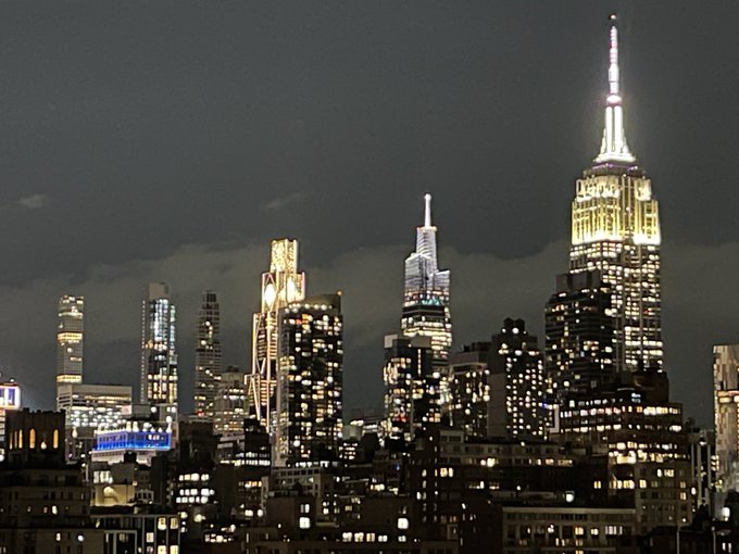 A nighttime photograph of the Manhattan skyline, featuring the brightly lit Empire State Building and One Vanderbilt (or similar modern skyscraper) against a cloudy dark sky.