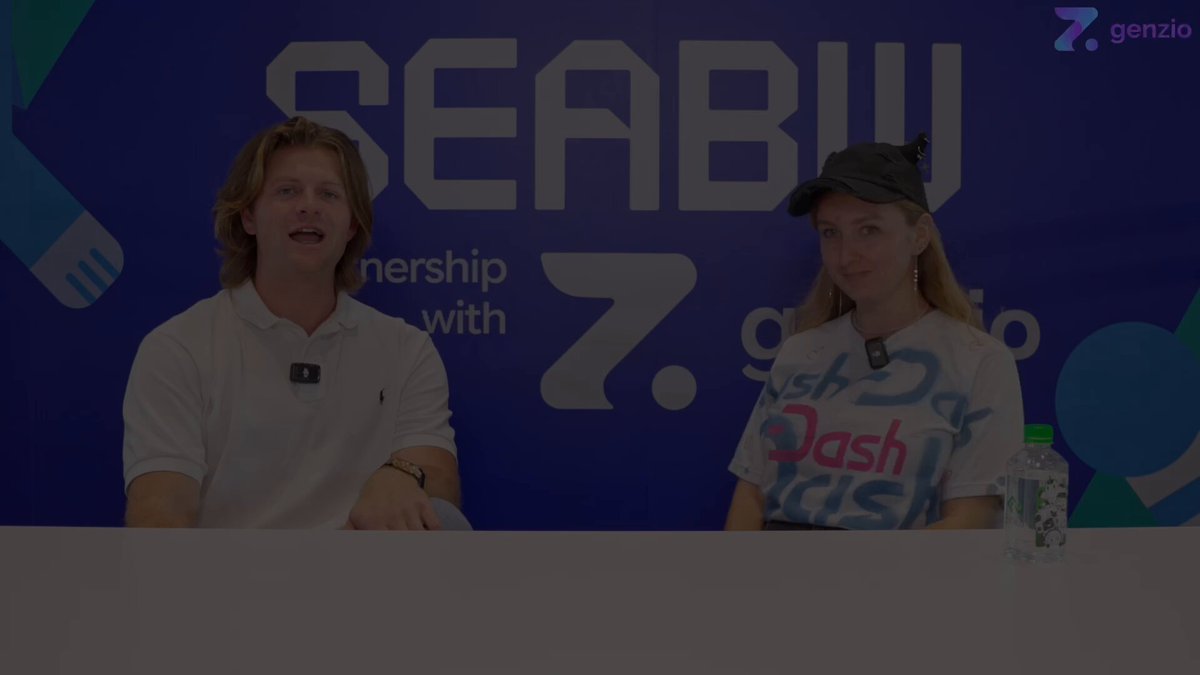 The image features a man and woman sitting behind a table with their backs facing each other at a conference or event. They appear to be engaged in conversation, possibly discussing various topics related to their experiences or interests. The background of the image includes a blue banner that reads "SEABWOfficial" and "@DariaChernozub". Additionally, there is a water bottle placed on the table between them.