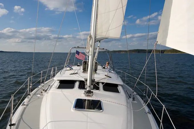 The image shows a white sailboat sailing across a body of water, with an American flag flying from its mast. The boat appears to be moving towards the right side of the frame. In the background, there are some buildings visible on the shoreline. The caption at the bottom of the photo reads "It's official I sold all my RWAs and bought all of Shiba and Barks crypto on a discount". This message seems to be related to the sailboat or its owner, possibly discussing their recent transactions in cryptocurrency.