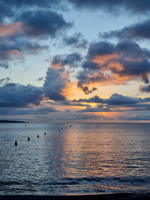 A serene sunset over the ocean with dramatic clouds in shades of orange, pink, and blue, and buoys visible on the water.