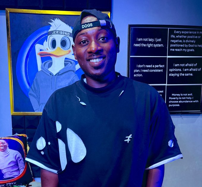 A young man wearing a "DOGS" cap smiles brightly in front of a framed painting of a duck wearing a "JERIBOND" cap and a motivational poster with quotes like "I am not lazy, I just need the right system." The image ties to the birthday celebration of the $DOGS token, with the cap and artwork reflecting its playful, meme-inspired branding.