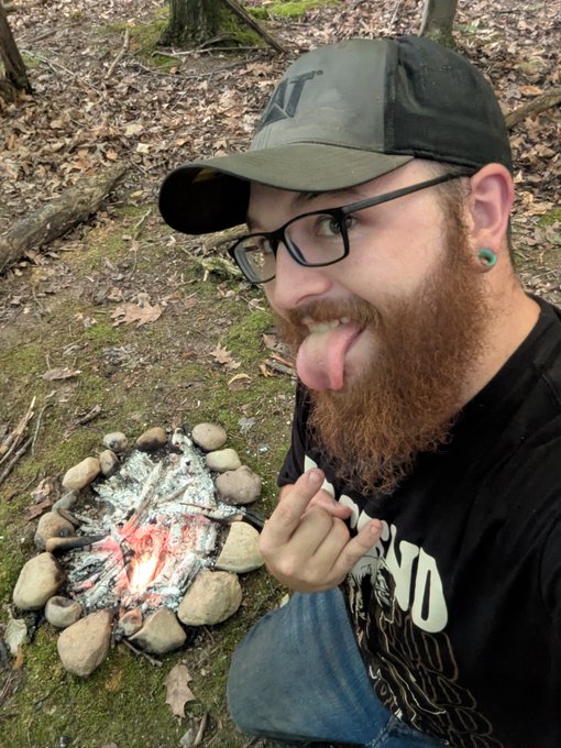 A man wearing a cap, glasses, and a band t-shirt sticks his tongue out at the camera while sitting next to a stone-ring campfire in a wooded area.