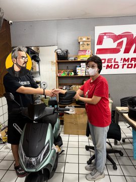 A photo of a man receiving a scooter from a dealership worker wearing a mask, with a 'DN' (Del' Max Motor) logo visible on the wall behind them.