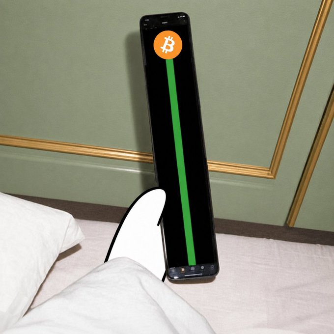 Smartphone displaying a Bitcoin logo and a green line/graph on a black screen, propped up against a green wall with gold trim on a bed with white pillows.