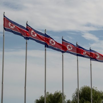 Five North Korean flags flutter against a cloudy backdrop, visually anchoring news that North Korea stole at least $2.8 billion in crypto between January 2024 and September 2025, increasingly laundering it through scam syndicates and organized crime networks.