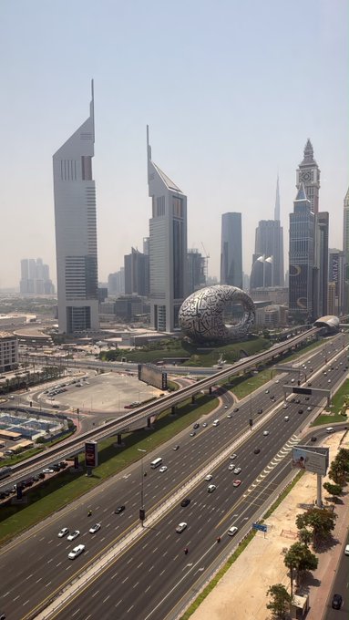 An aerial cityscape of Dubai featuring the Museum of the Future (the torus-shaped building), the Emirates Towers, and a multi-lane highway with light traffic.