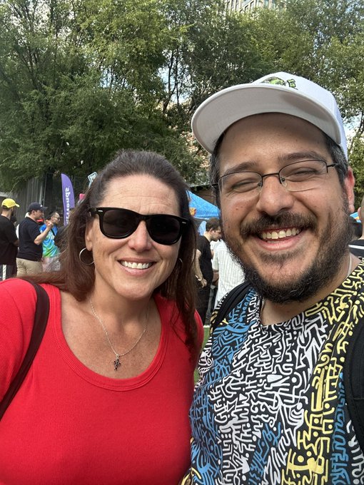 A selfie of a smiling couple outdoors at what appears to be a festival; the woman wears sunglasses and a red top, while the man wears glasses, a white cap, and a colorful patterned shirt.