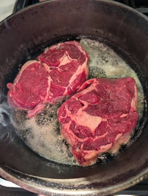 Overhead photo of two raw steaks cooking in a cast iron skillet with melted butter, beginning to sear and brown.