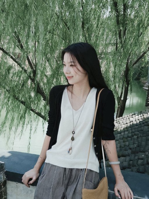 A young woman with long black hair wearing a white top, black cardigan, and grey pants stands by a railing near a willow tree and water.
