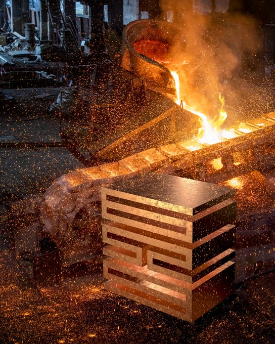 The image features a metal cube sitting next to a fire pit with sparks flying around it. The cube appears to be made of copper and has an interesting design. The fire pit seems to be used for melting down the metal, possibly creating a piece of art or a sculpture. The surrounding area is filled with various pieces of machinery and equipment, indicating that this space might be a workshop or a factory where such processes take place.