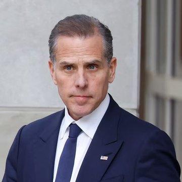 A middle-aged man with graying hair, wearing a dark navy suit, white shirt, and dark blue tie with a small American flag pin, looks directly at the camera with a serious expression.