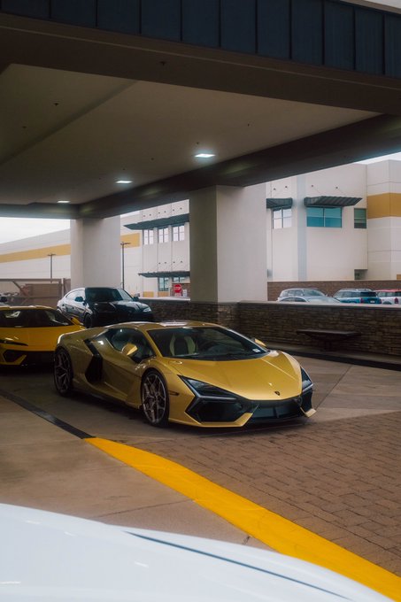 A gold Lamborghini Revuelto parked under an overhang, with another yellow sports car visible behind it in what appears to be a parking area.