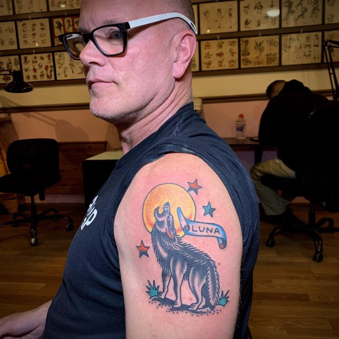 A man in glasses and a black tank top displays a colorful wolf tattoo with a 'LUNA' banner and stars on his upper arm, standing in what appears to be a tattoo parlor.