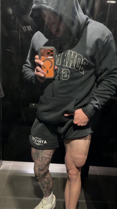 A man takes a mirror selfie in a dark gray "ATHLFT" hoodie and matching shorts, holding an orange phone case. The image reflects the post's message about physical health and fitness, showcasing a lean, muscular build with visible tattoos as evidence of disciplined health habits.