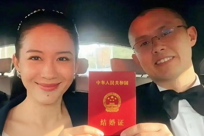 A smiling couple holds up a Chinese marriage certificate (中华人民共和国 结婚证) for the camera, apparently responding to skeptics who doubted their marital status by proudly displaying the official red booklet.