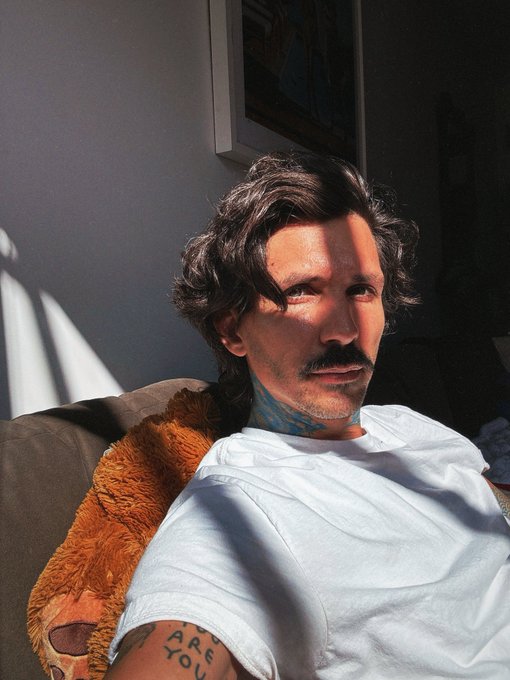 A selfie of a man with dark wavy hair and a mustache wearing a white shirt, sitting on a couch with a tattoo reading 'ARE YOU' on his arm.