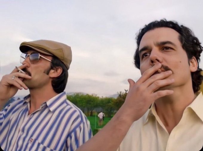 Two men smoke cigarettes while looking out over a scenic landscape, one wearing a striped shirt, sunglasses, and a flat cap, and the other in a cream-colored shirt with curly dark hair. The relaxed, contemplative pose of the men smoking fits the vibe of casually waiting for or reflecting on wins, matching the user's hopeful post about expecting more victories today and preparing to share cash with mutuals.