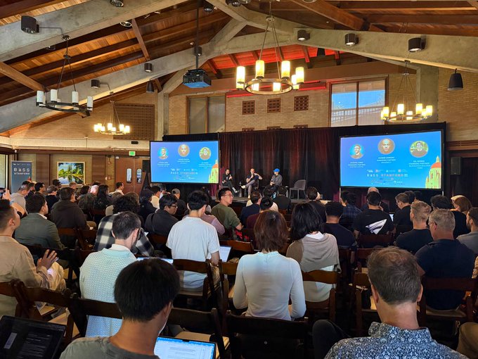 An audience seated in a large hall with a vaulted wooden ceiling, watching a panel discussion or presentation on stage featuring speakers seated between two large projection screens.