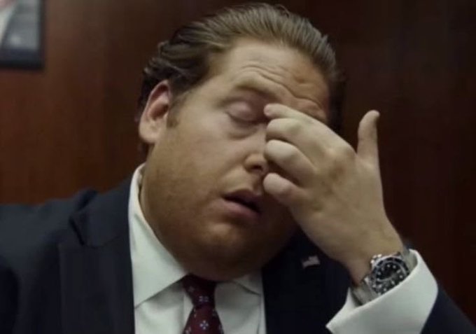 A man in a dark suit and red tie rubs his eyes and forehead in apparent frustration or fatigue, wearing a silver wristwatch.