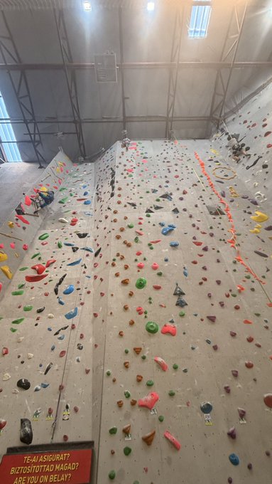 A wide view of an indoor rock climbing gym with multiple walls covered in colorful climbing holds, featuring a sign that reads 'TE-AI ASIGURATI BIZTOSITOTTAD NAGAD? ARE YOU ON BELAY?'