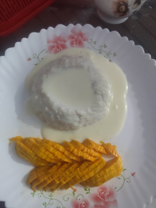 A white floral-patterned plate holds a creamy rice porridge or pudding drizzled with milk, served alongside crispy golden plantain or banana slices, presented as a hearty dinner.