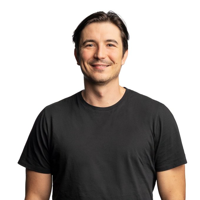 A portrait photo of a smiling man with short dark hair wearing a black t-shirt against a plain white background.