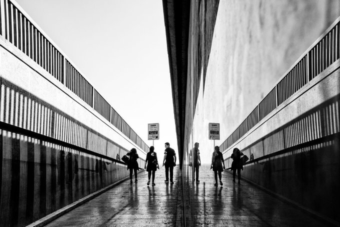 A black and white photograph of silhouetted figures walking through a modern architectural corridor with reflective walls creating a mirror effect, showing signs that read 'NO TODOS LOS DIAS' and 'NO TODOS'.