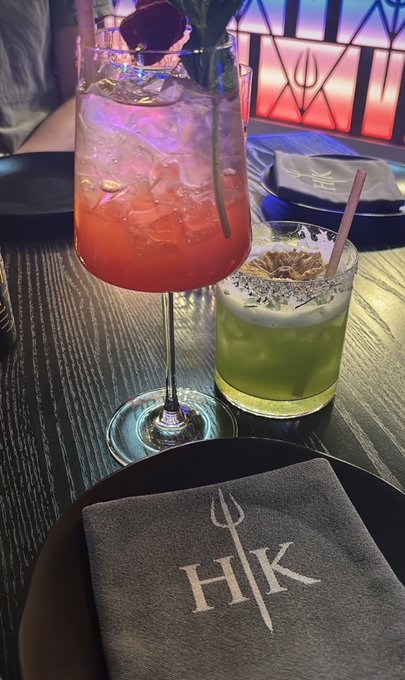 The image shows a dining table with two drinks, one pink and one green, placed on plates. There are also napkins on the table that have the word "Hk" written on them. The background of the photo features a window with red and blue lights, adding a vibrant touch to the scene.