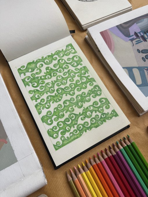 An open sketchbook displays a green geometric pattern of swirling shapes drawn in colored pencil, with an assortment of colored pencils arranged nearby on a wooden surface.