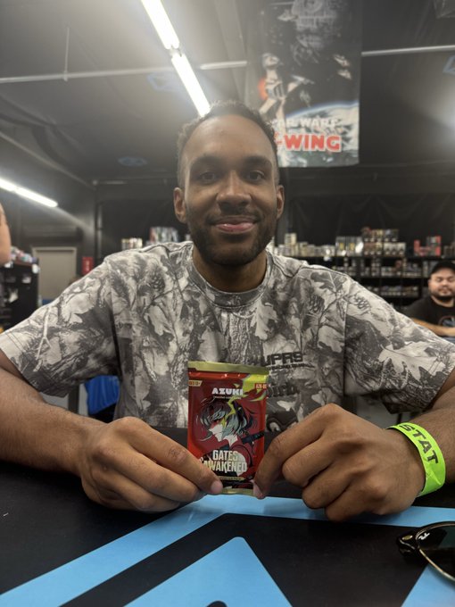 A man smiles at the camera while holding an Azuki "Gates of Unawakening" trading card pack, with an "Azuki WING" sign visible in the background, perfectly matching the post about ripping some Azuki packs.