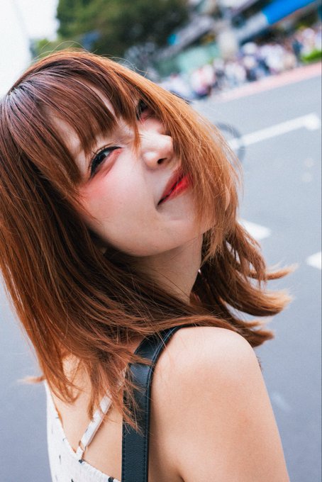 A young woman with shoulder-length reddish-brown hair and bangs looks over her shoulder at the camera while standing on a paved street, wearing a white top with a black strap.
